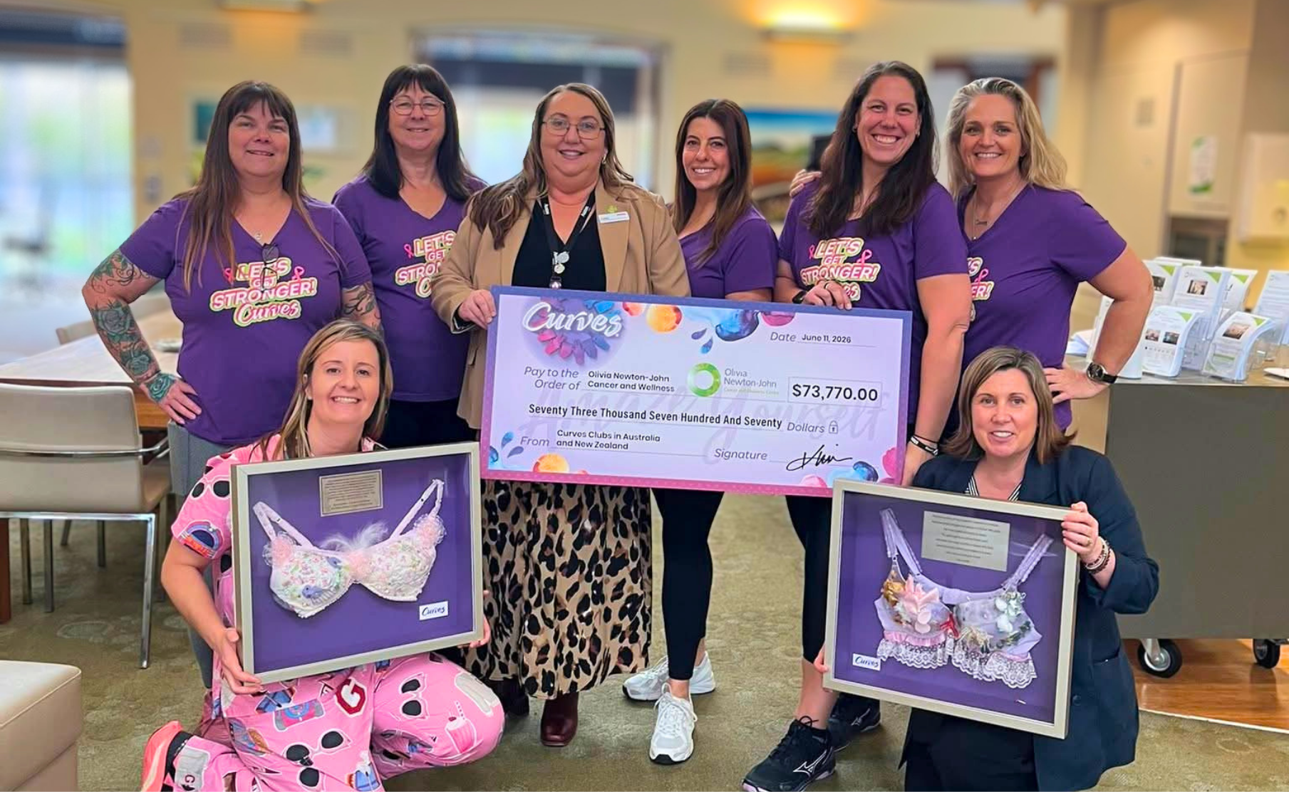 Seven members of the Curves Australia and New Zealand team smiling and holding a giant fundraising cheque of ,770 for the Olivia Newton-John Cancer and Wellness Centre, flanked by two framed, decorated contest bras.