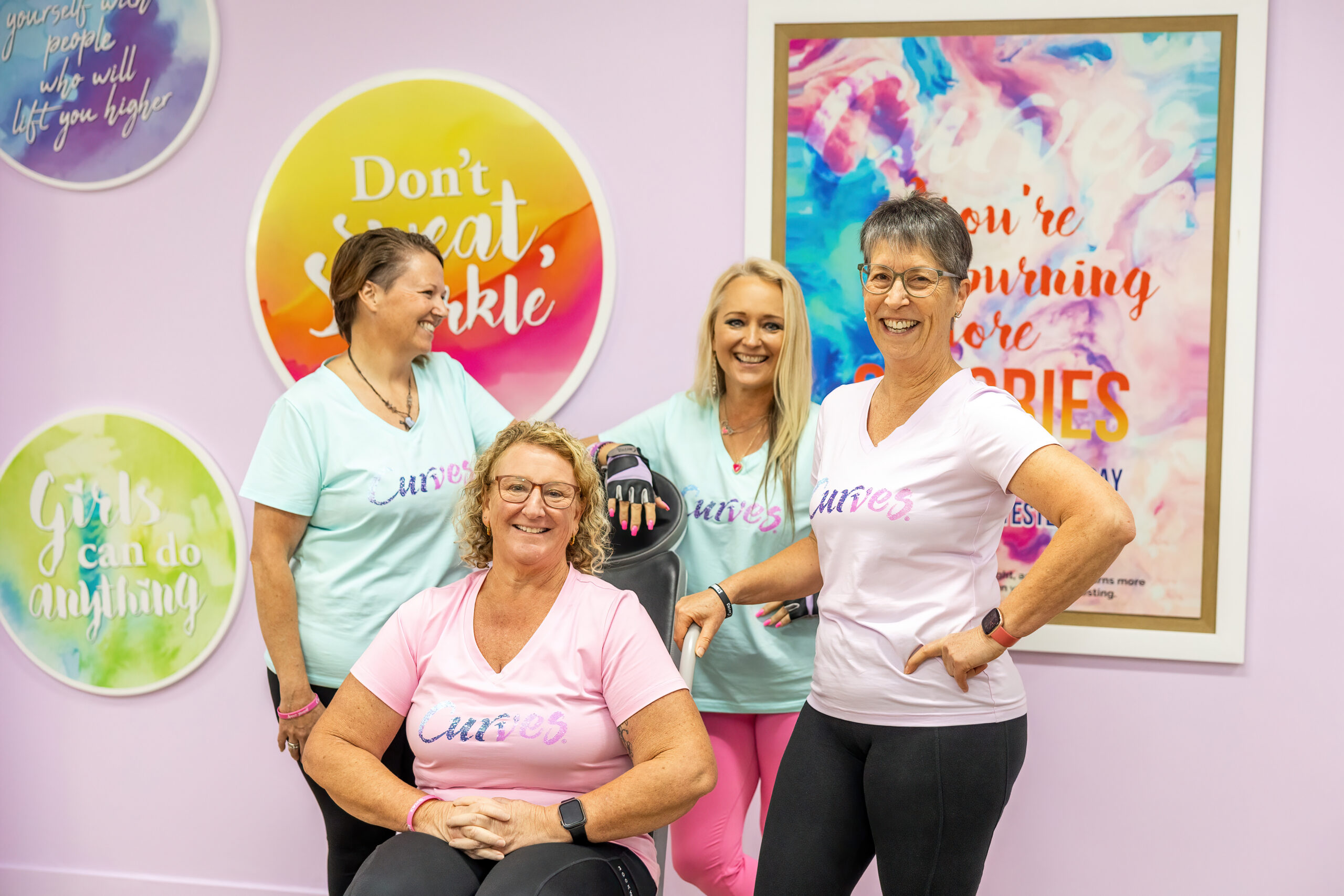 A multi-generational group of smiling women and Kim McQueen wearing Curves shirts posing together inside a brightly decorated women's strength training circuit.