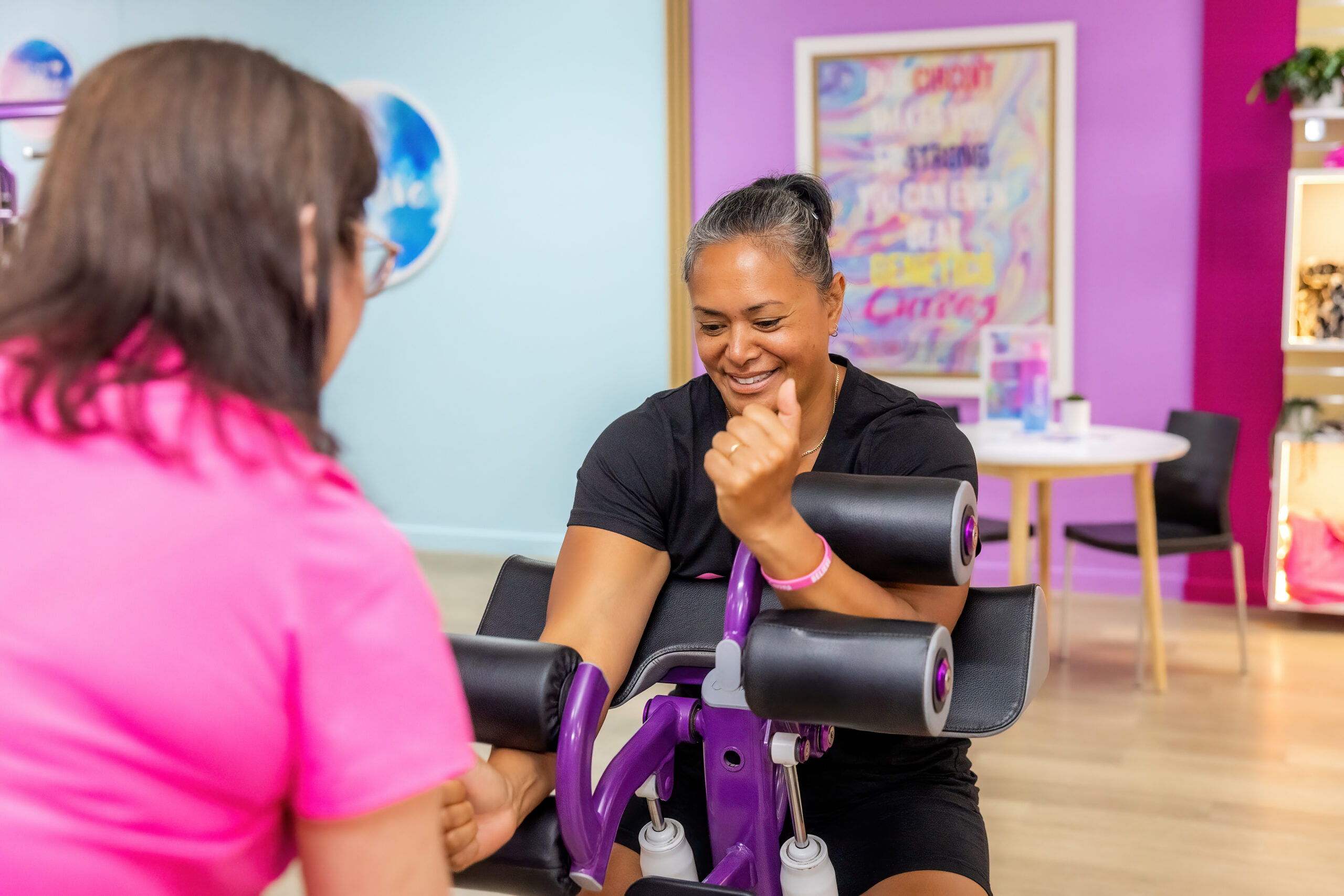 A smiling woman completing a workout on a purple hydraulic chest and back machine with the guidance of a Curves gym coach.
