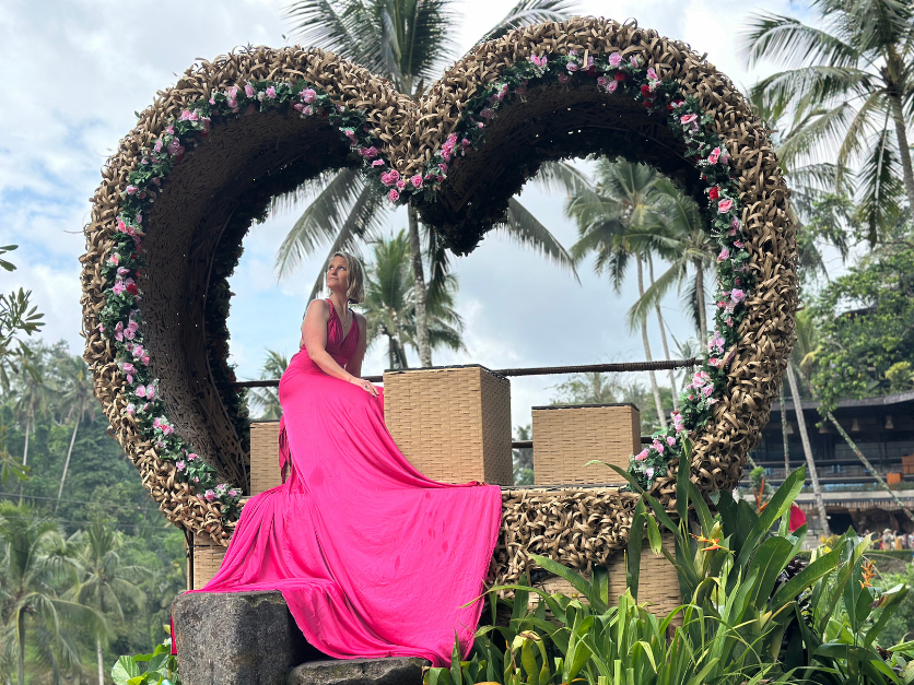 A woman in a flowing magenta dress sits smiling inside a large floral heart structure during the Curves Bali Retreat in Ubud.