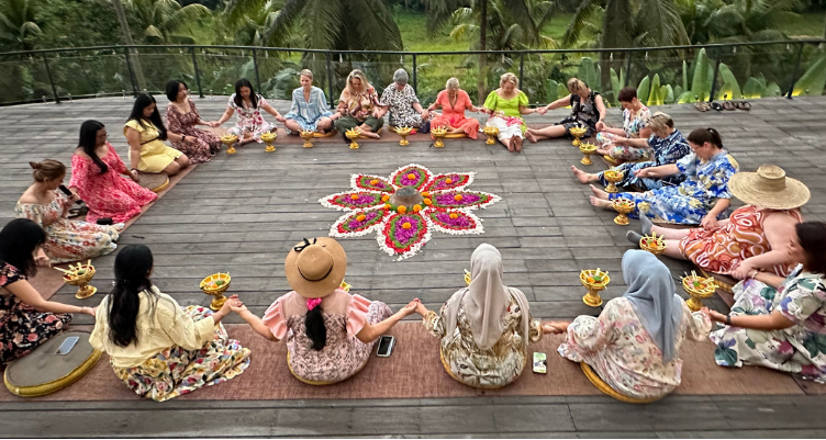 A diverse group of women holding hands in a large, supportive circle around a vibrant flower mandala ceremony during the Curves Bali Retreat at Soulshine Ubud.