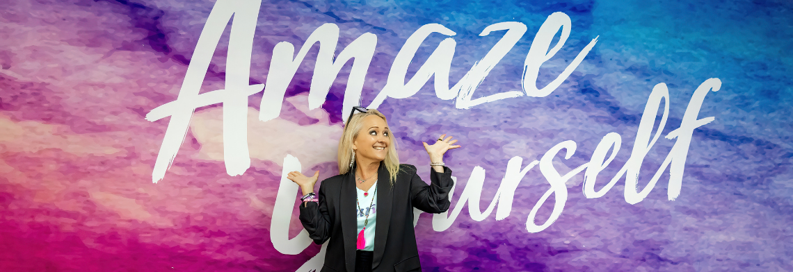 Curves CEO Kim McQueen smiling playfully in front of a purple and blue watercolor wall that reads "Amaze Yourself."