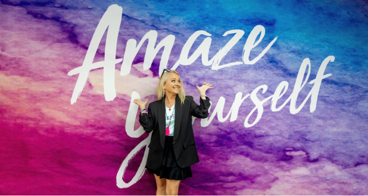 Curves CEO Kim McQueen smiling playfully in front of a purple and blue watercolor wall that reads "Amaze Yourself."
